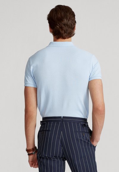 Mand iført lyseblå kortærmet polo og marineblå bukser med tynde striber, stående med ryggen til kameraet mod en grå baggrund.