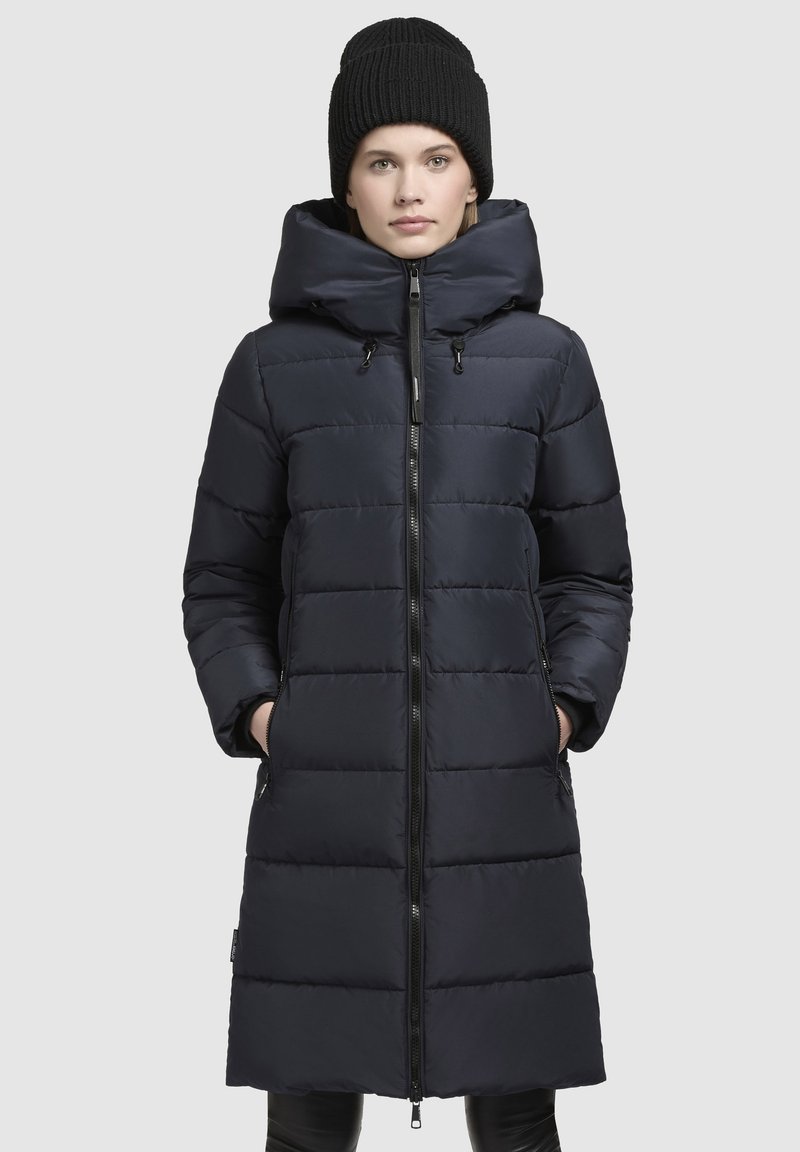 Navy padded long coat with a hood, featuring a front zipper, two side pockets, and a quilted design. Paired with a black knit beanie.