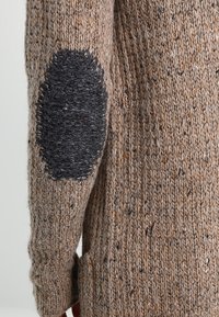 Stickad tröja i en brun och beige blandning med en texturerad yta, med en stor mörkgrå armbågsjacka. Sidoflik för ökad komfort.