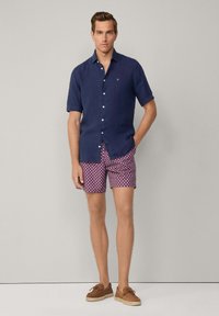 Marineblaues Kurzarmhemd mit Knöpfen, kombiniert mit rosa Shorts, die ein kleines weißes Sternmuster aufweisen. Braune Slipper runden den Look ab.