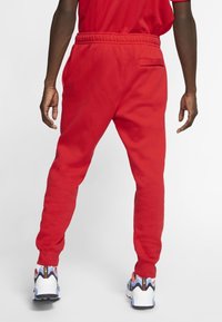 Red sweatpants featuring an elastic waistband and a back pocket, paired with multicolored athletic shoes on a neutral background.