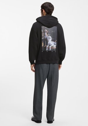 Man met een zwarte hoodie met afbeelding van twee honden op een stoel in de bibliotheek, gecombineerd met grijze broek en zwarte schoenen, staat met zijn rug naar de camera.
