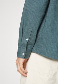 Camicia in tessuto a spina di pesce verde scuro con maniche lunghe, polsini con bottoni e un dettaglio della tasca, abbinata a pantaloni di colore chiaro.