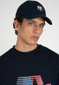 Joven con gorra azul oscuro de NY, camisa azul marino con gráfico rojo y azul, y un tatuaje vertical en el cuello contra un fondo claro y liso.