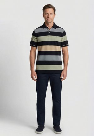 Mann steht und trägt ein kurzärmliges Poloshirt mit breiten horizontalen schwarzen, grauen, grünen und beigen Streifen, dunkle Hose und dunkle Slipper.