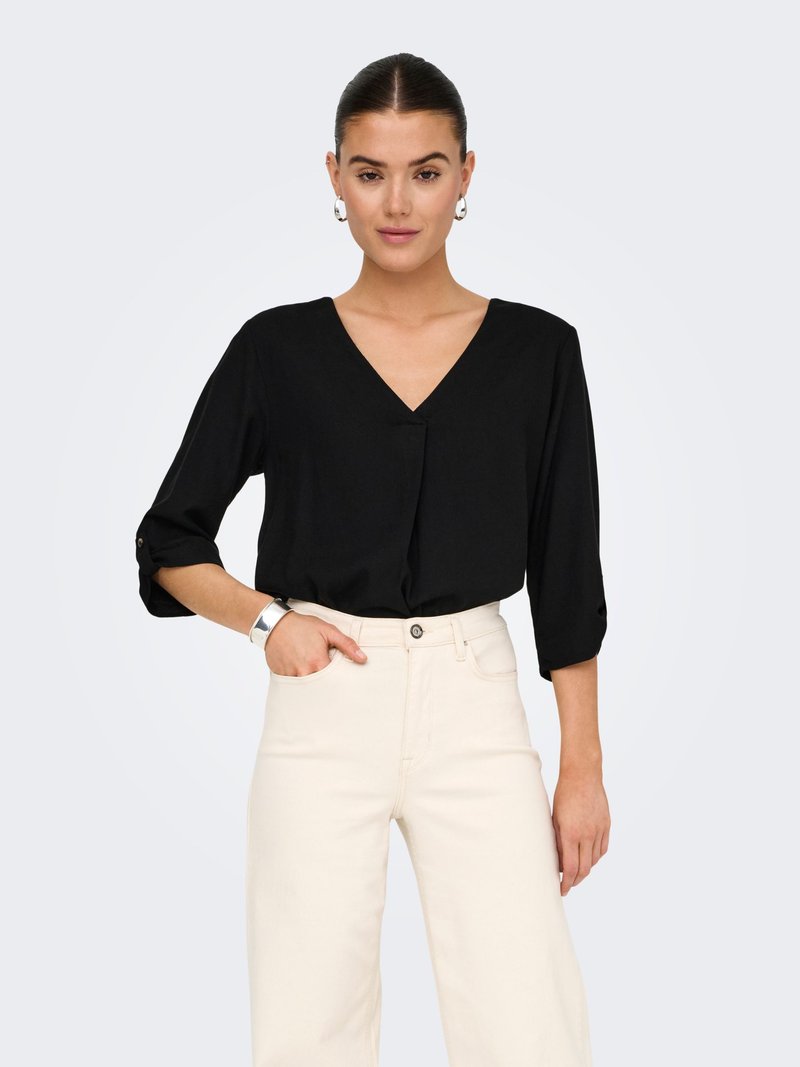 Femme portant un haut noir à col en V avec manches 3/4 et pantalon taille haute écru, posant avec une main dans la poche sur fond blanc.