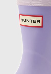 Purppuranvärinen kuminen sadekenkä, jossa on teksturoitu vaaleanpunainen yläosa ja suorakulmainen punainen ja kerma valkoinen etiketti, jossa lukee "HUNTER". Sileä pintaviimeistely.