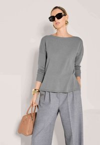 Frau in einem übergroßen grauen Pullover und grauen, gefalteten Hosen, die eine beige geflochtene Handtasche hält, große schwarze Sonnenbrille und goldene Accessoires trägt.