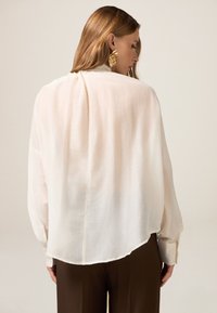 Blusa ligera de color crema con un corte holgado, mangas largas y un dobladillo curvado. Presenta una textura suave y un diseño sencillo.