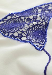Tissu délicat en dentelle bleue avec des motifs floraux bordés de petites perles, posé sur une surface blanche lisse.