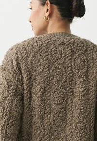 Brązowy sweter o fakturze z miękkiego materiału przypominającego boucle, z okrągłymi wzorami, prążkowanymi mankietami oraz prostym fasonem.