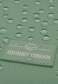 Zöld, textúrált felület vízcseppekkel, rajta egy téglalap alakú, fehér betűkkel "JOHNNY URBAN" feliratozott, dombornyomott gumi folt.