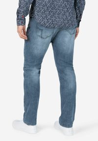 Lichtblauwe spijkerbroek met een slim fit, subtiele vervaging en twee achterzakken, gecombineerd met witte sneakers en een donker shirt met een patroon.