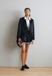 Blazer nero oversize, camicia bianca con colletto, gonna mini in tessuto, e sneakers nere. Capelli lisci, in piedi contro un muro semplice.