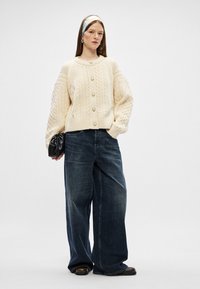 Cárdigan de punto trenzado color crema con botones de perla, combinado con unos vaqueros oscuros y holgados. El look se completa con un bolso de mano negro.