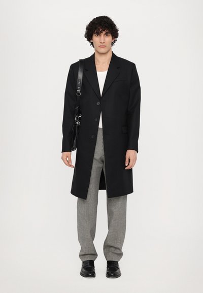 Schwarzer langer Mantel mit knöpfbarem Vorderteil, kombiniert mit grauen karierter Hose und schwarzen Schuhen. Das Modell hält eine schwarze Schultertasche.