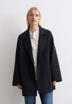 Black oversized coat with a double-breasted design, large lapels, and wide cuffs. Worn over a white polka dot shirt and blue jeans.