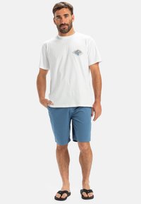 Vitt bomullströja med en liten färgglad grafik på bröstet, tillsammans med blå shorts. Svarta flip-flops fulländar outfiten.