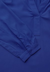 Camisa azul de manga larga, diseño de cuello en V, textura fruncida en el collar y puños con botones. Acabado de tela suave.