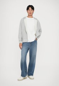 Joven de pie que lleva puesta una sudadera con cremallera gris, una camiseta blanca, jeans azules holgados y zapatillas blancas frente a un fondo blanco.
