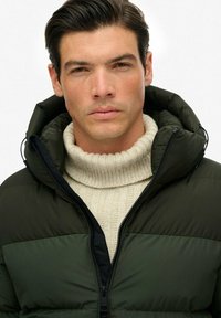 Grøn pufferjakke med hætte, med sort lynlås og syninger. Båret over en cremefarvet turtleneck sweater med en glat overflade og struktureret pasform.