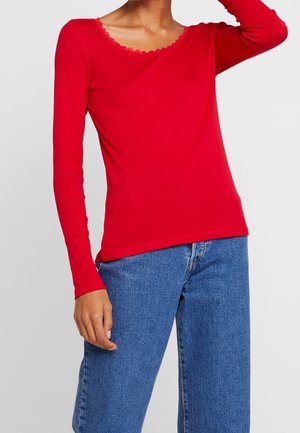 Kobieta w dopasowanym czerwonym topie z długim rękawem i wycięciem w falowanym kształcie oraz niebieskich, wysokich dżinsach na tle jednolitego tła.