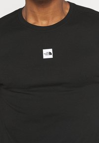 Černé bavlněné tričko s kulatým výstřihem. Na hrudi se nachází bílé čtvercové logo s textem a zakřivenými grafickými prvky. Hladký povrch.