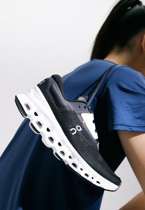 Black knit running shoe with white sole and distinct holes, featuring a prominent logo and clean lines. The shoe is held by a person in a blue top.