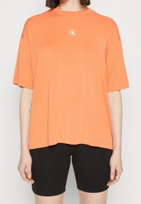 Camiseta de algodón naranja con cuello redondo y mangas cortas, que presenta un logo blanco "cK" en el pecho. Combinada con pantalones cortos negros.