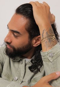 Goldene Mehrstrang-Armbänder an einem tätowierten Handgelenk; Model trägt eine hellgrüne Jeansjacke mit metallenen Druckknöpfen; lange dunkle Haare.