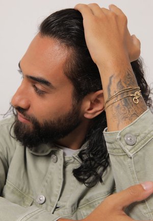 Guld flertrådet armbånd på et tatoveret håndled; model iført en lysegrøn denimjakke med metaltryklåse; langt mørkt hår.