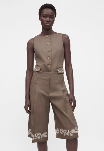 Γυναίκα που φοράει αμάνικη καφέ ολόσωμη φόρμα με κουμπιά στο μπροστινό μέρος, τσέπες με καπάκι και λευκή κεντητή λεπτομέρεια παϊέτες στο τελείωμα, σε απλό φόντο.