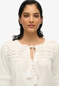 Blusa bianca con tessuto testurizzato, dettagli in pizzo e scollo a cravatta. Presenta piccoli bottoni e nappine all'estremità della cravatta.