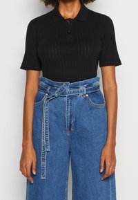 Person som bär en kortärmad svart ribbad polo skjorta ihop med högmidjade blå denimjeans med ett knytbälte.