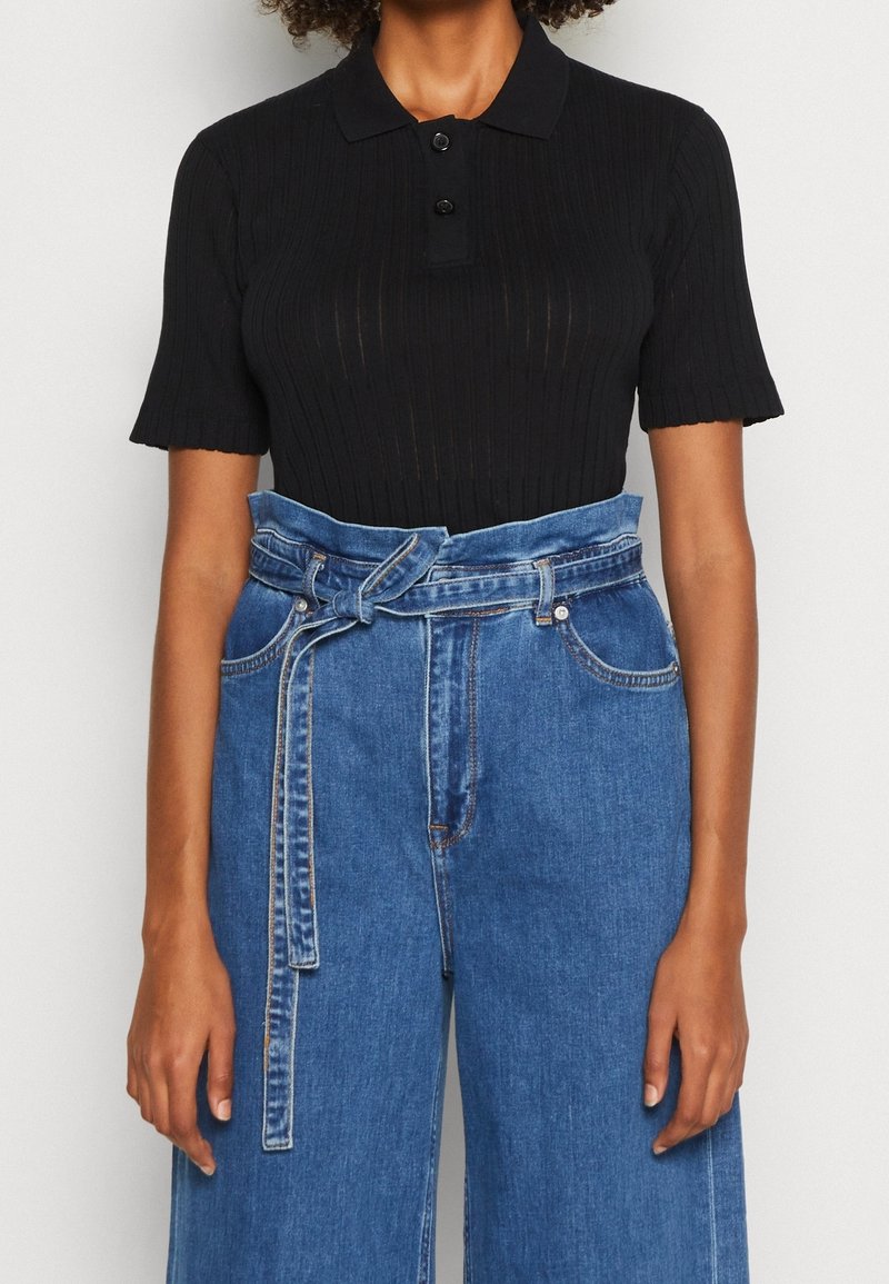 Person som bär en kortärmad svart ribbad polo skjorta ihop med högmidjade blå denimjeans med ett knytbälte.