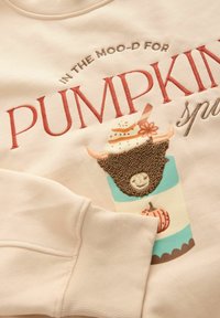 Lichtbeige sweatshirt met geborduurde tekst, een beker met een glimlachend gezicht, een gekruld personage en decoratieve pompoenaccenten.