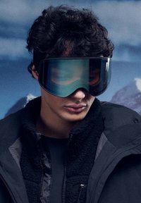 Skibrille mit einer breiten, gewölbten Linse in dunklen Blau- und Grüntönen, mit einem schlanken Design. Getragen zu einer schwarzen, mit Fleecefutter versehenen Jacke.