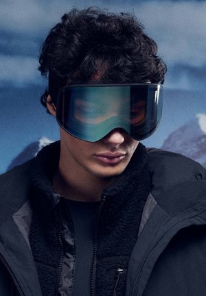 Suusaprillid laia, kõverdatud läätsedega tumesinistes ja rohelistes toonides, mis omavad sujuvat disaini. Kandmises must fliisvoodriga jope.