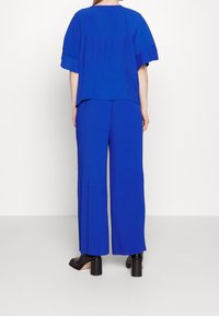 Osoba stojąca, ubrana w jasnoniebieską, prążkowaną bluzkę z krótkim rękawem oraz szerokie spodnie, połączona z czarnymi botkami na obcasie, na białym tle.