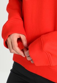 Pullover rosso con polsini e orlo a coste. In primo piano, mani che regolano il polsino, mostrando la texture del tessuto e il colore vivace.