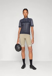 Dviračių apranga: tamsiai mėlynas trumpomis rankovėmis džersis ir šviesiai smėlio spalvos šortai su tamsiomis detalėmis. Rankoje laikoma juoda šalmas, matomos juodos bateliai ir kojinės.