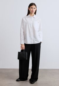 Λευκό πουκάμισο με κουμπιά και τσέπη στο στήθος, σε συνδυασμό με μαύρο παντελόνι φαρδιάς γραμμής. Το μοντέλο κρατά μια μικρή μαύρη υφασμάτινη τσάντα.