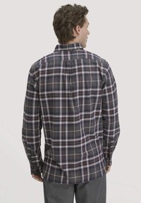 Man die met zijn rug naar de camera staat, gekleed in een donkerblauw en wit geblokt overhemd met lange mouwen en grijze broek, tegen een effen lichte achtergrond.