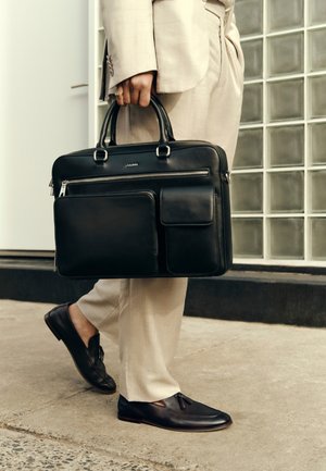 Isik, kes kannab beeži ülikonda ja musti loafer-tüüpi kingi, kannab musta nahast portfelli mitme lukuga sektsiooniga kõnniteel.