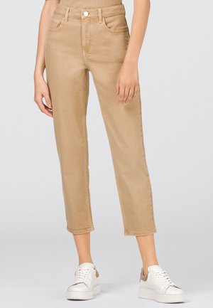 Beiget korkeavyötäröiset suoralahkeiset housut yhdistettynä valkoisiin korollisiin lenkkareihin, joita käyttää seisova henkilö yksinkertaisen taustan edessä.