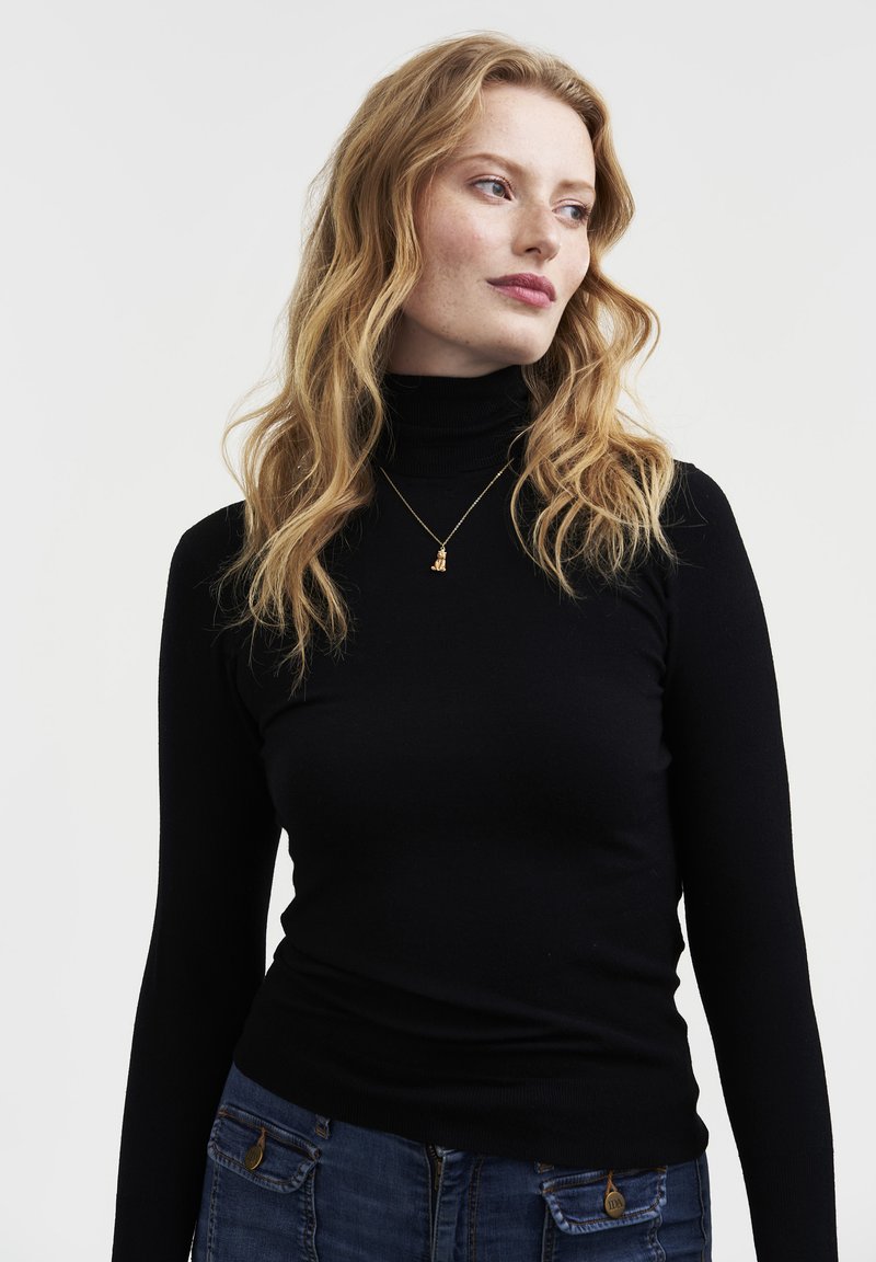 Kobieta o falowanych blond włosach, ubrana w czarny golf, niebieskie dżinsy z kieszeniami oraz mały złoty naszyjnik z zawieszką, patrząca w prawo.