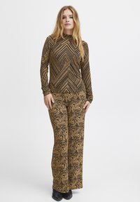 Langärmliges Oberteil und weit geschnittene Hose aus einem goldenen und schwarzen gemusterten Stoff mit geometrischen und floralen Designs, strukturiert für visuelles Interesse.