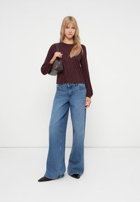 Bordeauxrotes Langarmoberteil mit gerippter Textur, kombiniert mit weit geschnittener blauer Jeans. In der Hand ein kleiner grauer Handtasche; schwarze spitze Schuhe an den Füßen.