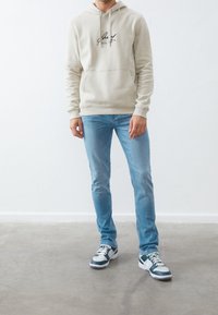 Sudadera beige con logo, bolsillo tipo canguro frontal; jeans ajustados de mezclilla azul claro; zapatillas negras y blancas con detalles en azul teal. Fondo liso.
