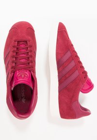 Röda mockasneakers med rosa accenter, med tre mörkrosa ränder, runda snören och en vit gummisula.